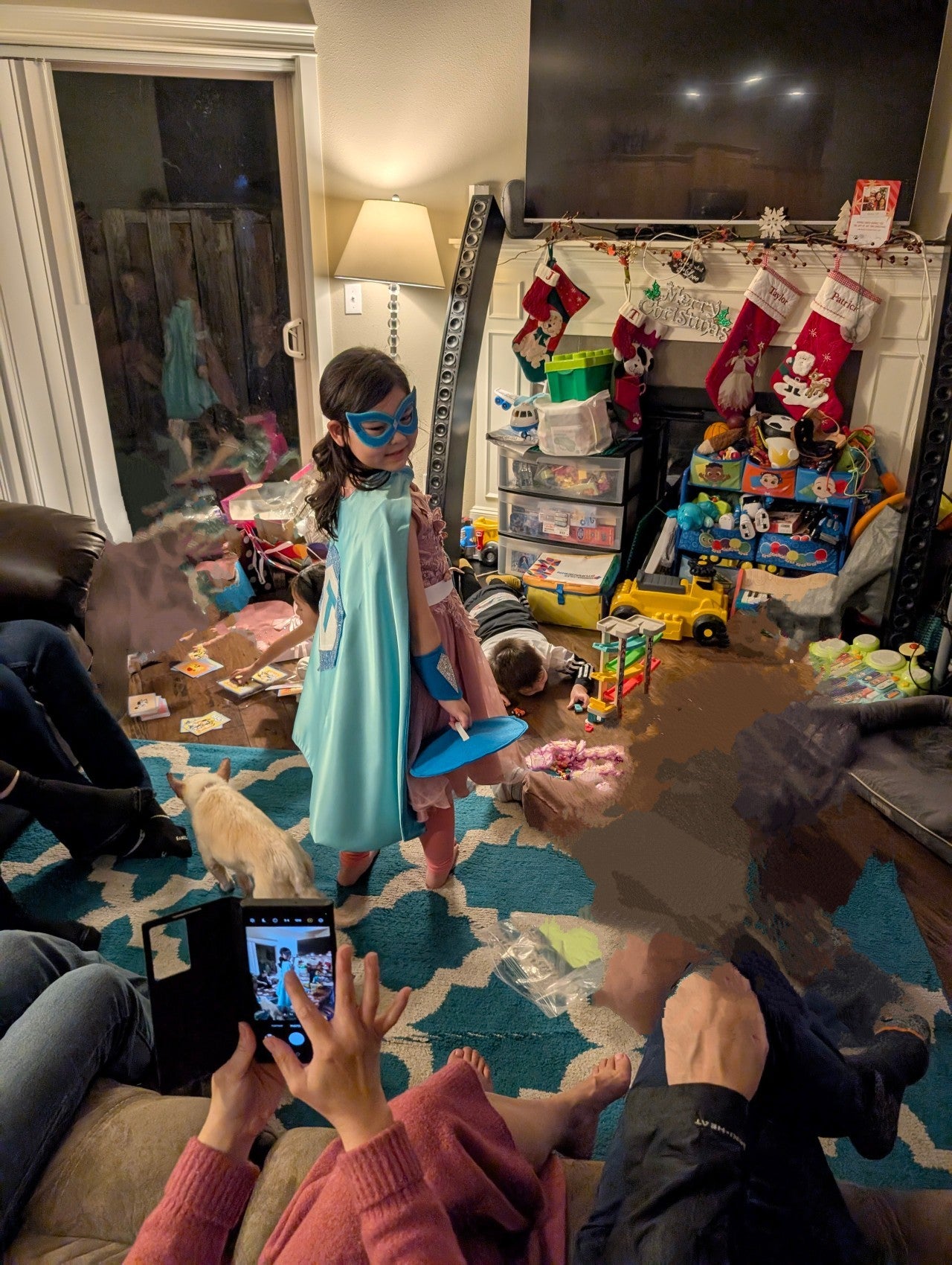 living room on Christmas morning and kids surrounded by toys. Girl is proudly modeling her blue satin super hero costume to family.