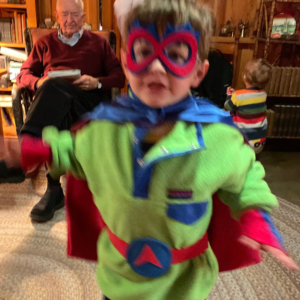 Active toddler in superhero costume with cape and mask in a room with an older person.