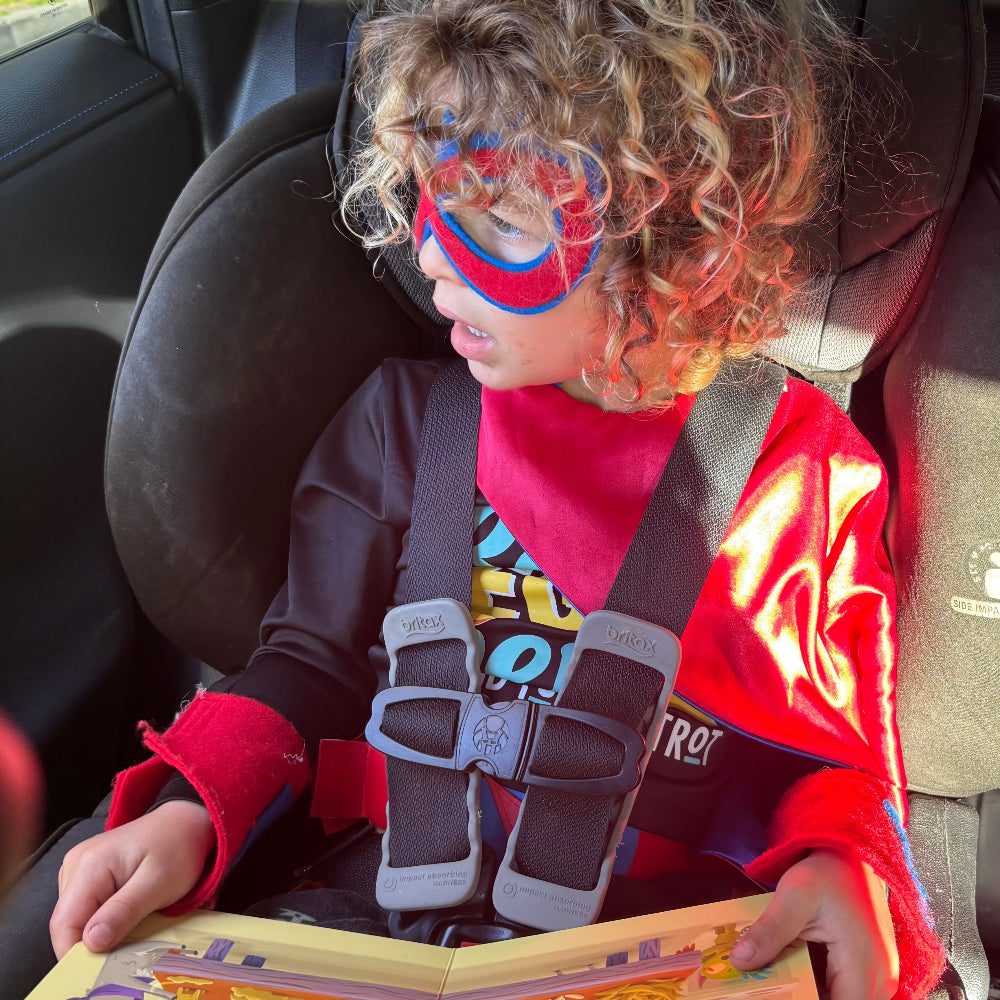 Child wearing a red superhero mask and costume sitting in a car seat with a book.