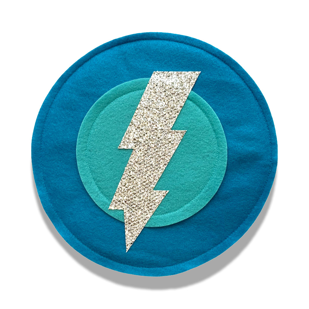 A dark aqua colored felt shield with a silver bolt design in the center, approximately 10 inches in diameter, with a soft elastic handle on the backside.