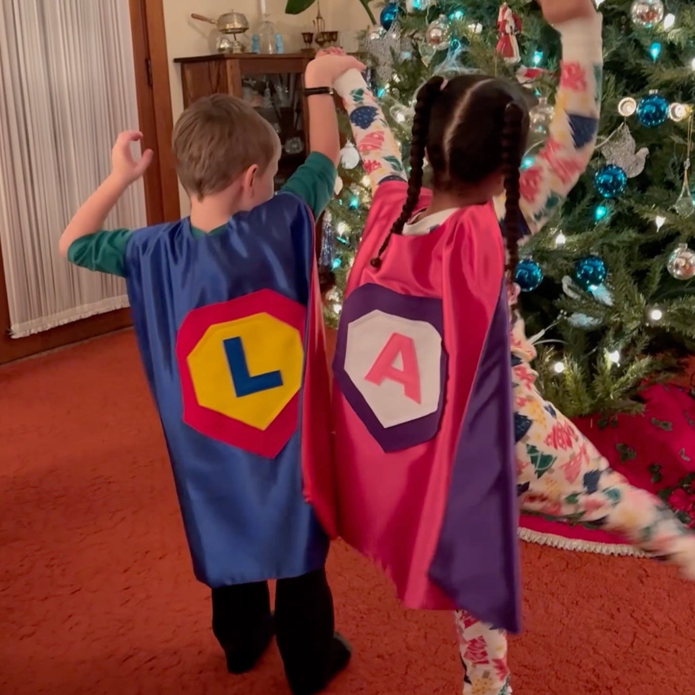 Happy boy and girl excited on Christmas to get Creative Capes superhero costume. Blue and Pink satin capes have the child's initial on the emblem