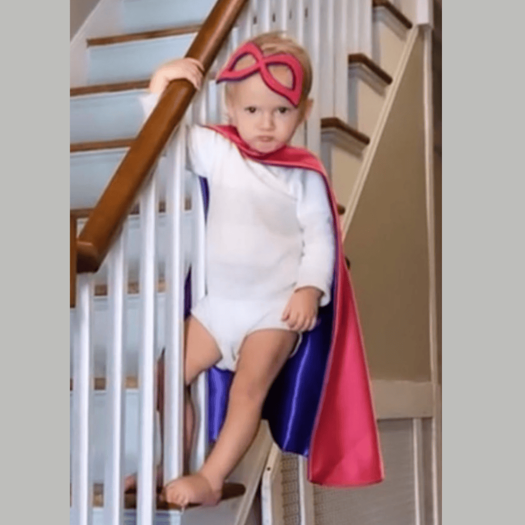 Child in pink and purple superhero costume with cape and eye mask on staircase. She is the winner of Creative Capes photo contest