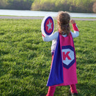 girl empowered in purple and pink personalized superhero costume set, superhero cape featuring the child's first initial, dress-up costume - lined cape, shield, mask, belt, and wrist cuffs, handmade by Creative Capes in USA to inspire imaginative play and confidence, Play activity for healthy childhood, no screen time