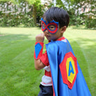 boy in blue, red and yellow superhero costume set, featuring the child's first initial on the super hero cape, quality dress-up costume kit includes a lined satin cape, shield, mask, belt, and wrist cuffs, handmade by Creative Capes in USA to inspire imaginative play and confidence. Play without screens creates a healthy childhood.