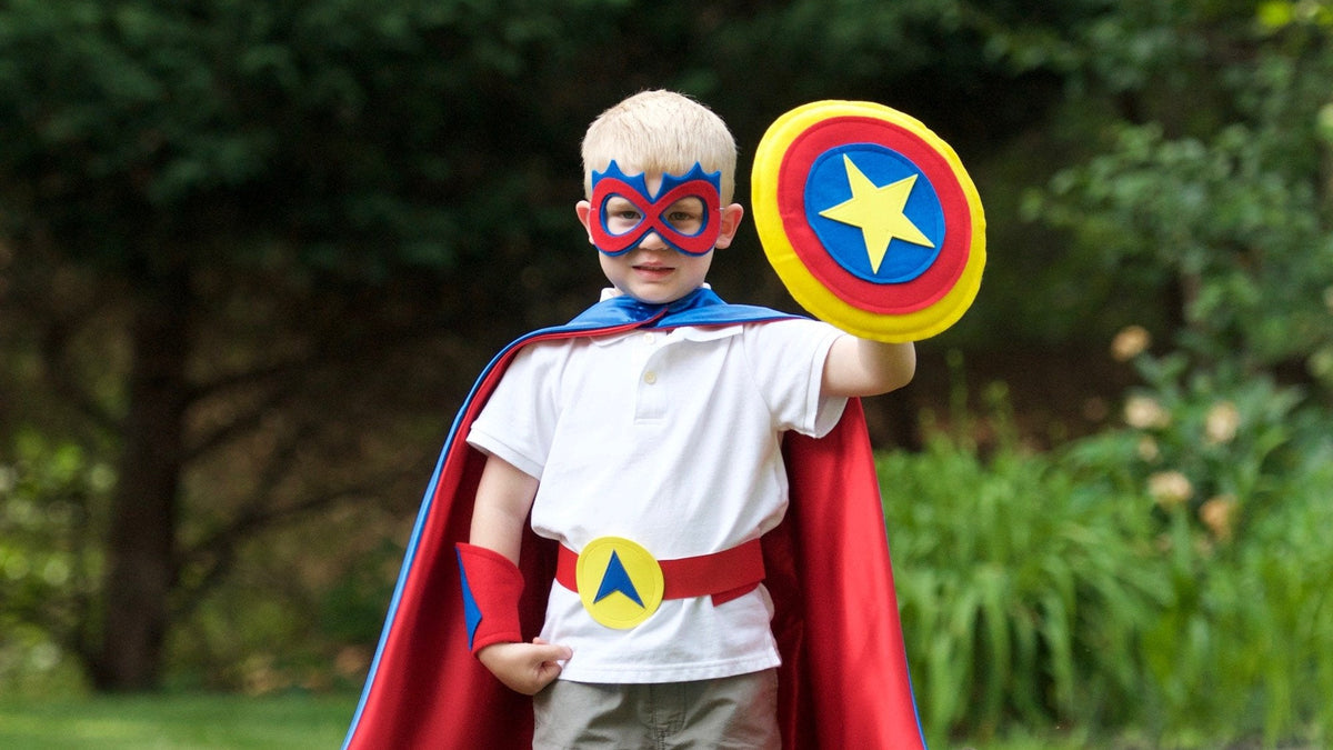 boy in blue, red and yellow superhero costume set, featuring the child's first initial on the super hero cape, quality dress-up costume kit includes a lined satin cape, shield, mask, belt, and wrist cuffs, handmade by Creative Capes in USA to inspire imaginative play and confidence. Play without screens creates a healthy childhood.
