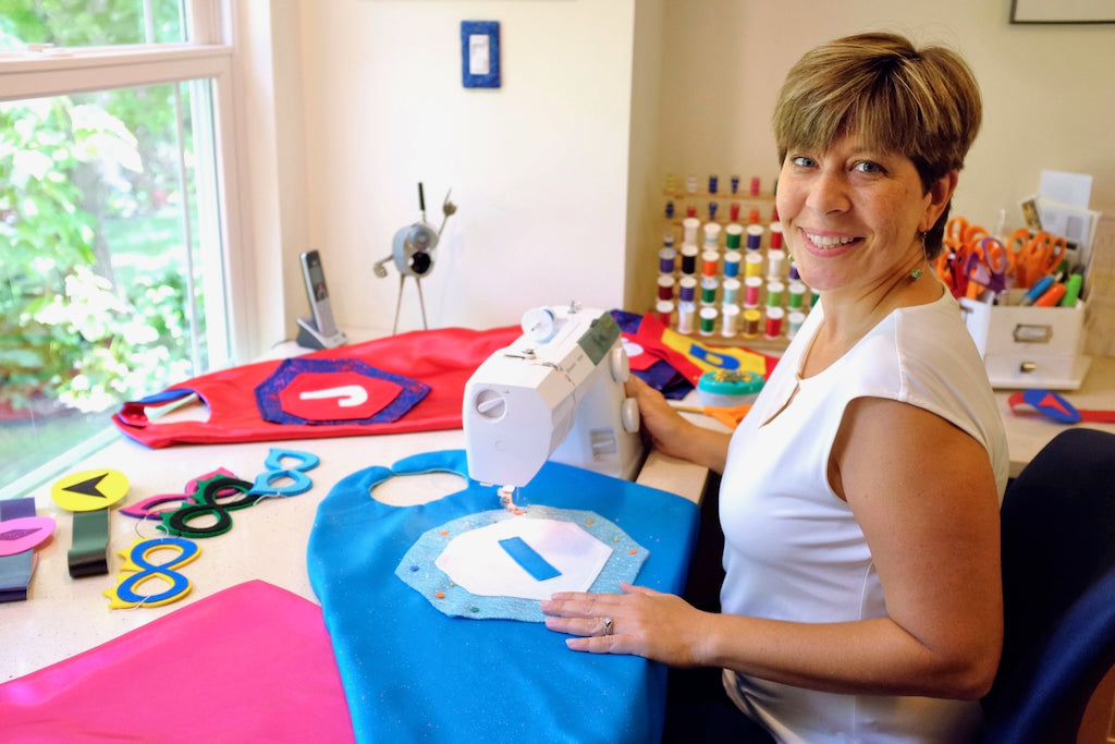 woman at sewing machine surrounded by satin custom superhero capes, Holly is the Artist and Founder of CreativeCapes.com