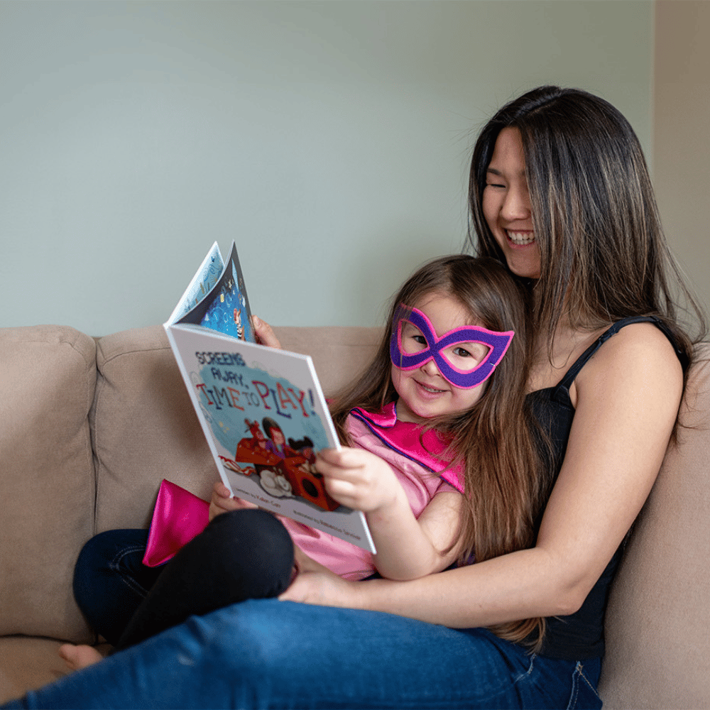 Mother and child reading a book together on a couch, with the child wearing a superhero mask and cape by CreativeCapes.com