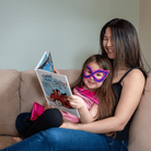 Mother and child reading a book together on a couch, with the child wearing a superhero mask and cape by CreativeCapes.com