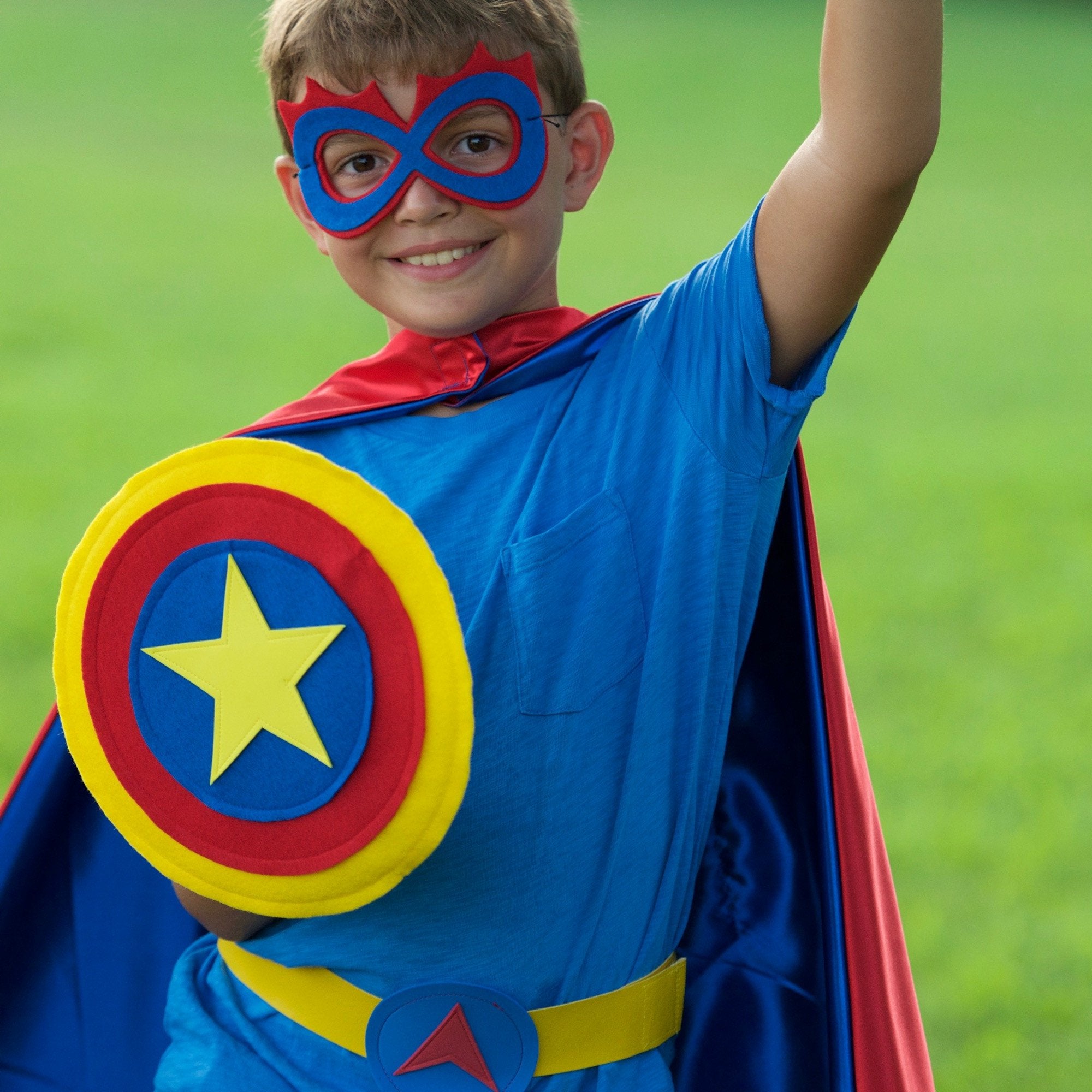 Active boy in Red, blue and yellow personalized superhero costume set, dress-up costume kit has a lined satin cape, shield, mask, belt, and wrist cuffs, handmade by Creative Capes in USA, gift to inspire imaginative play and confidence. Play without screens